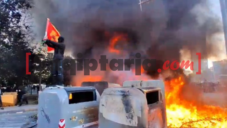 Protestuesi i maskuar hipën mbi koshat që digjen, valëvit flamurin e UÇK-së
